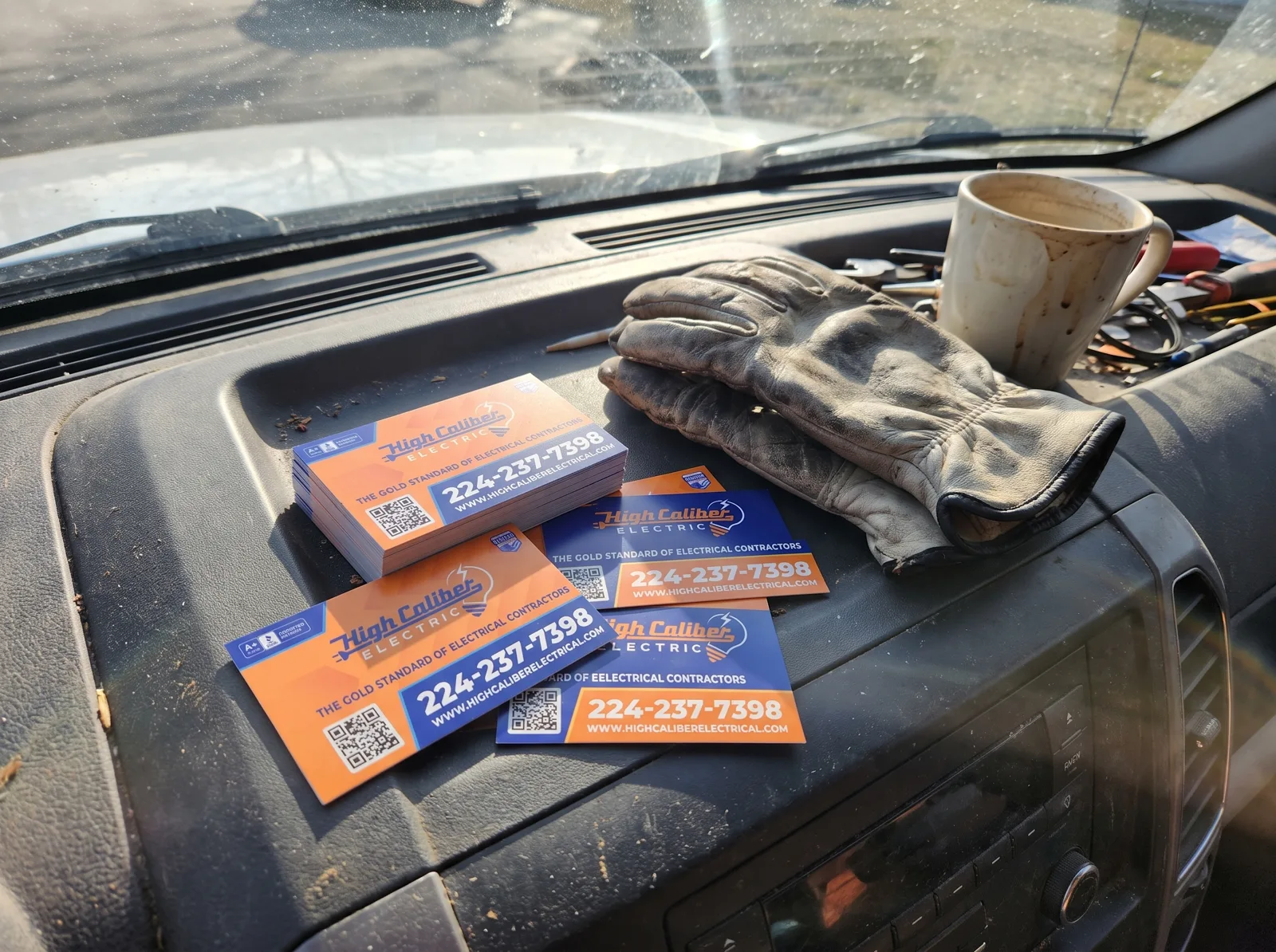 High Caliber Electric business cards on a dusty van dashboard with work gloves.