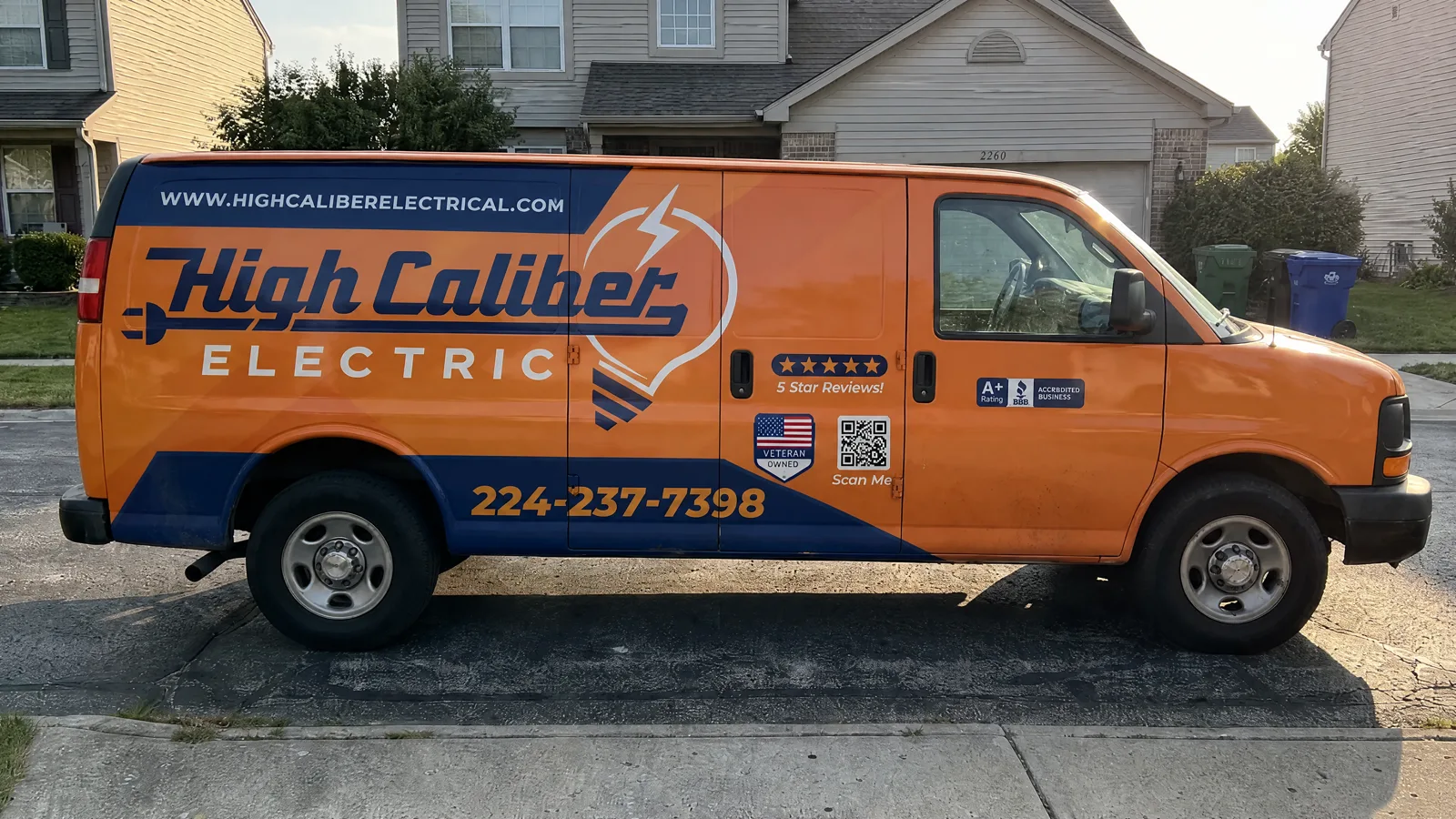 High Caliber Electric Chevy Express on a suburban street. Orange and navy wrap, High Caliber ELECTRIC, phone 224-237-7398, VETERAN OWNED shield on the sliding door.