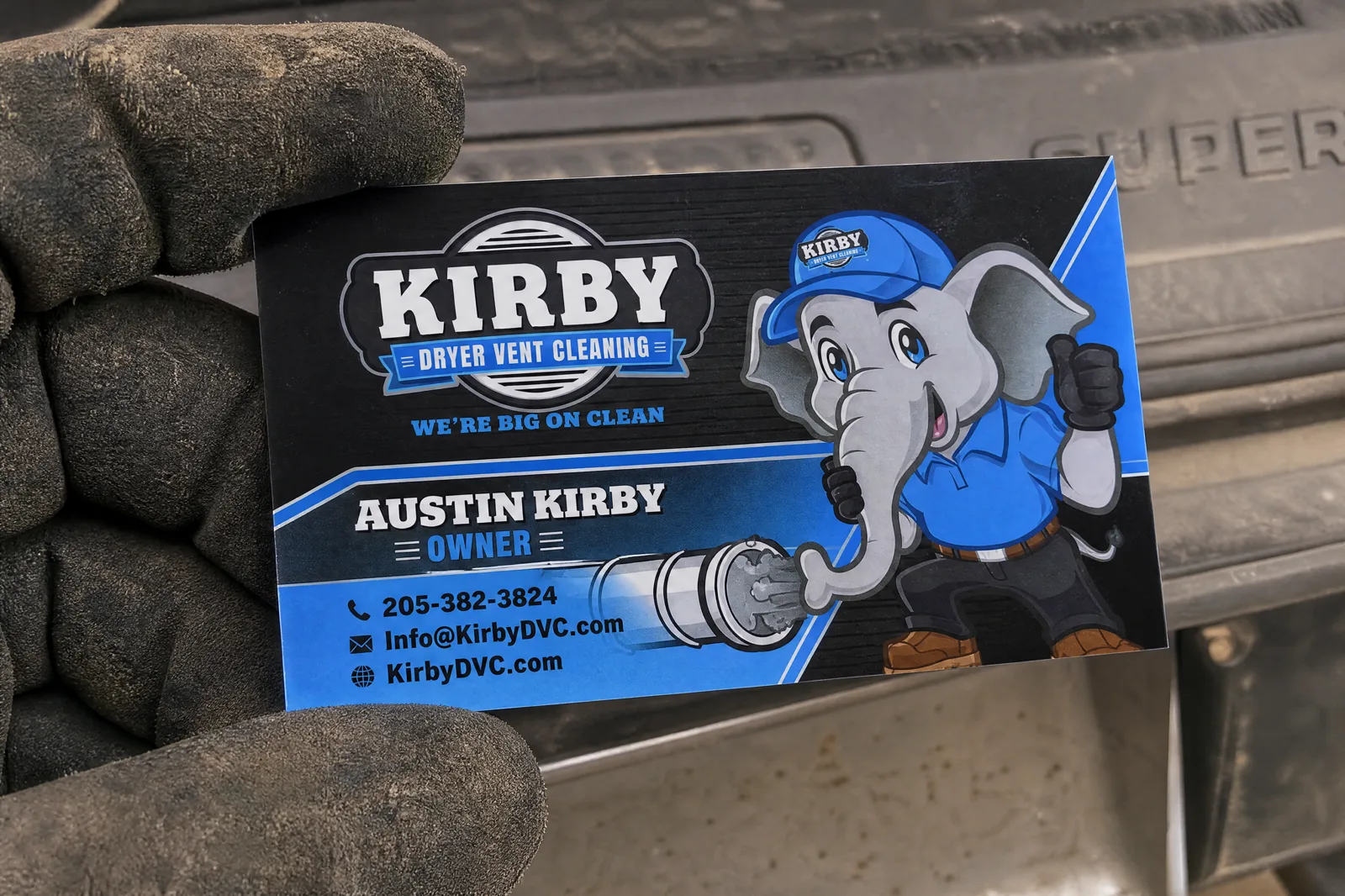 Kirby Dryer Vent Cleaning business card with elephant mascot, Austin Kirby owner, 205-382-3824 and KirbyDVC.com, held in a work-gloved hand.